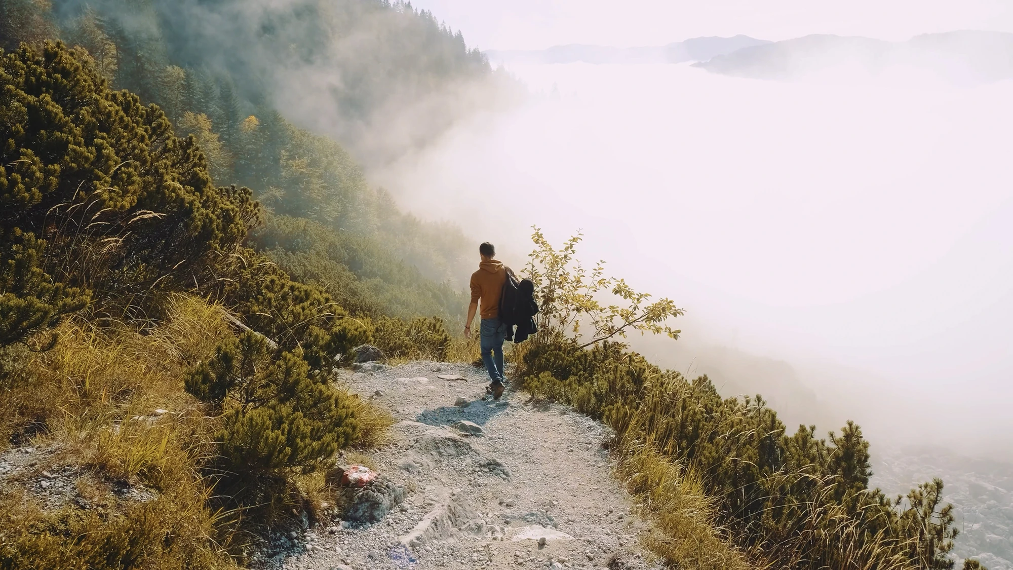 A hiker in an orange sweatshirt walks a gravel trail along a misty mountainside above a sea of clouds