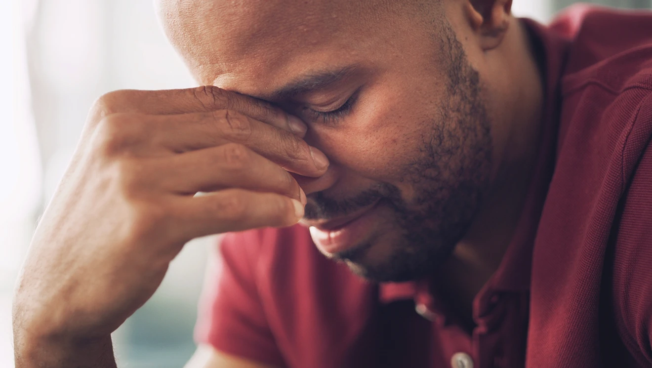 A man in a red polo shirt closes his eyes and presses his fingers to the bridge of his nose, looking weary