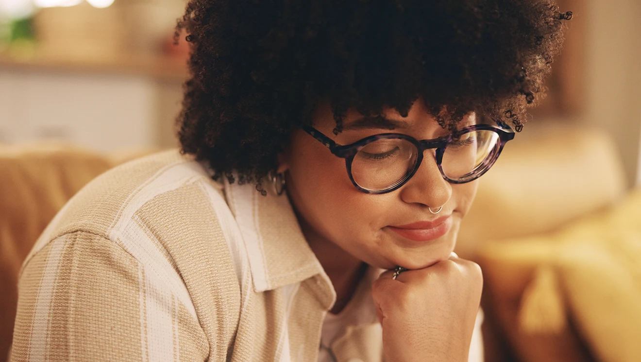 A person with dark curly hair and round glasses looks down thoughtfully, resting their chin on their hand in warm lamplight