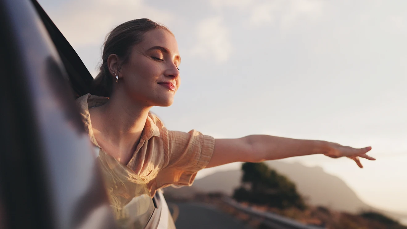 A young woman leans out of a car window at sunset with her eyes closed and her arm stretched out into the open air
