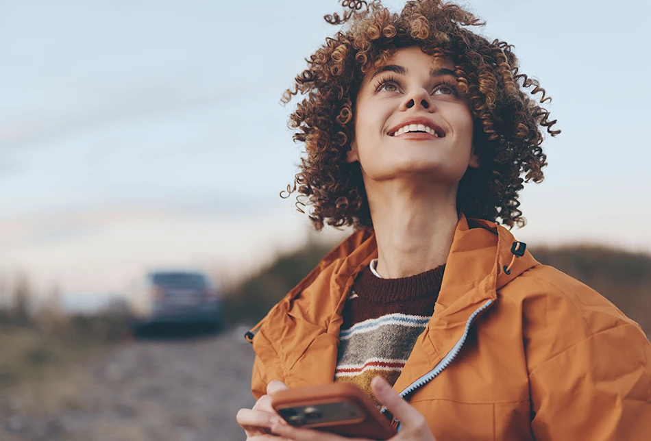 A young woman with curly hair looks up with a bright smile, holding her phone outdoors at dusk