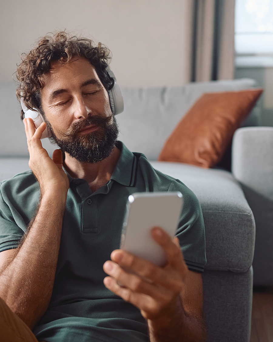 A man with curly hair and a beard sits against a sofa with his eyes closed, one hand on his headphones, holding a phone