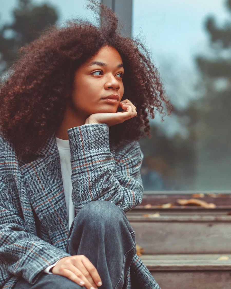 A young woman with curly hair sits on wooden steps, her chin resting on her hand, gazing thoughtfully to one side