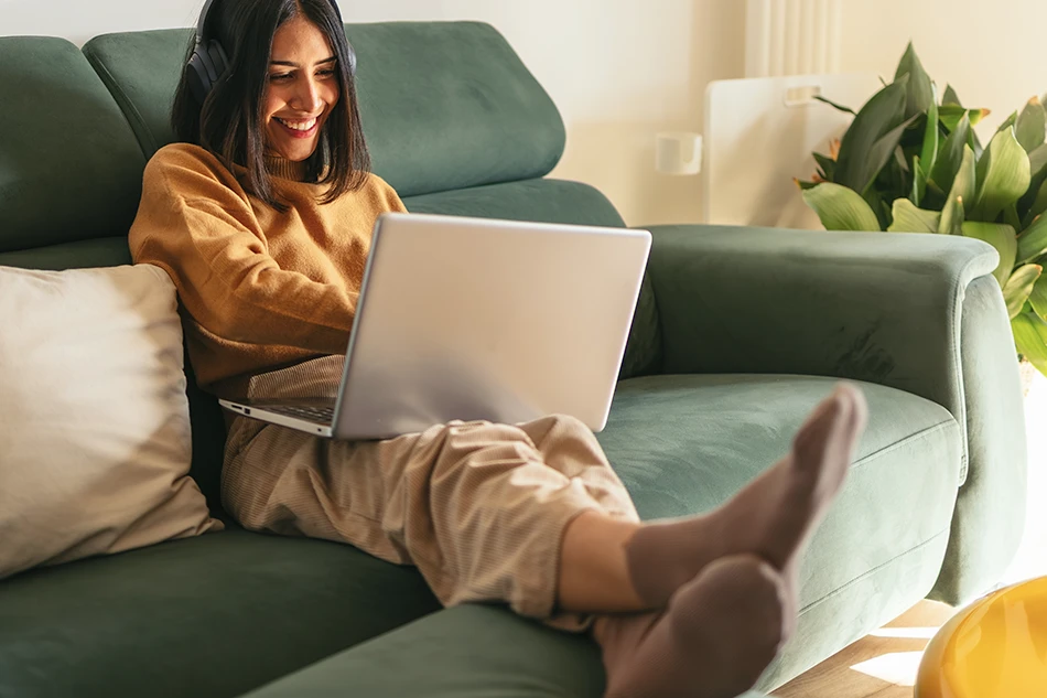 A smiling woman wearing headphones relaxes on a green sofa with a laptop on her lap and her feet up